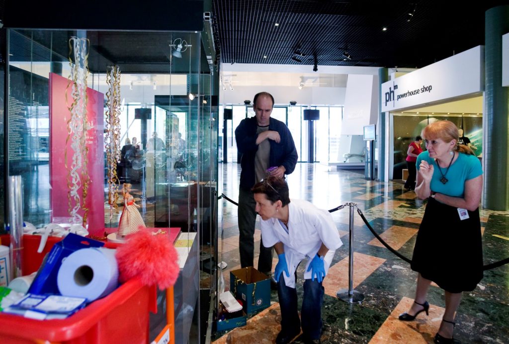 Three people are standing around a display case in the foyer of the Museum. They are looking at a Barbie doll wearing a long gold and white striped evening dress with an orange sash and long white gloves. The doll is standing in the centre of a small display case from which numerous lengths of white and gold curling ribbon cascade down either side of her from the top. A woman wearing a lab coat and blue latex gloves bobs down to look inside the display case. A trolley with cleaning equipment and a feather duster can be seen on the left. In the background can be seen a sign which reads 'Powerhouse Shop'. 